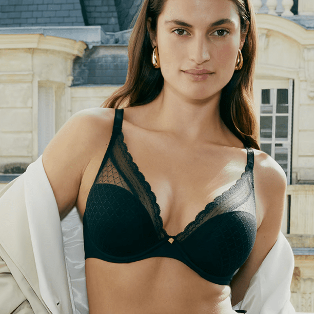 Woman wearing a black lace plunge bra with scalloped trim and moulded cups, styled with a light jacket draped off the shoulders, standing outdoors in front of a stone building.