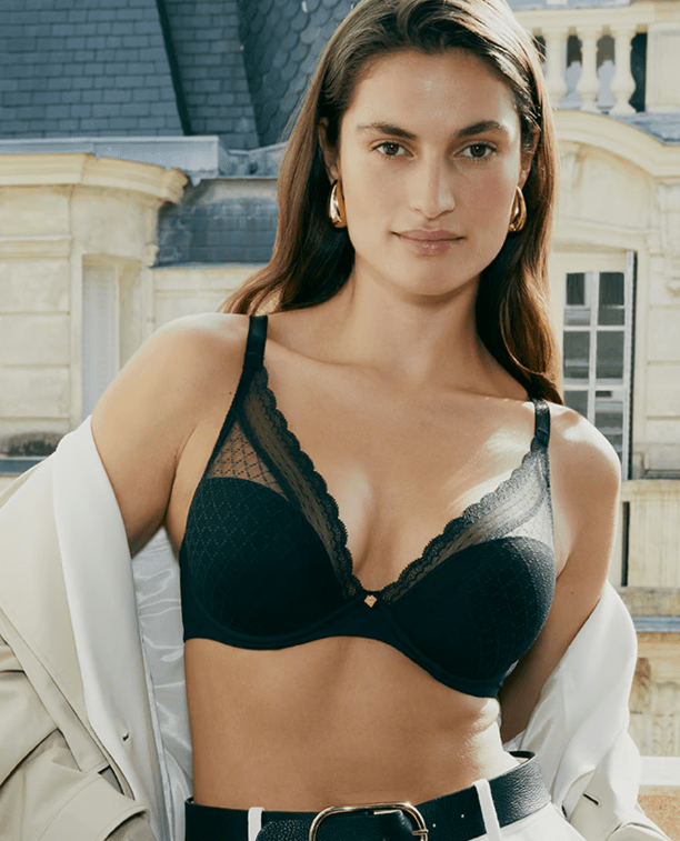 Woman wearing a black lace plunge bra with scalloped trim and moulded cups, styled with a light jacket draped off the shoulders, standing outdoors in front of a stone building.