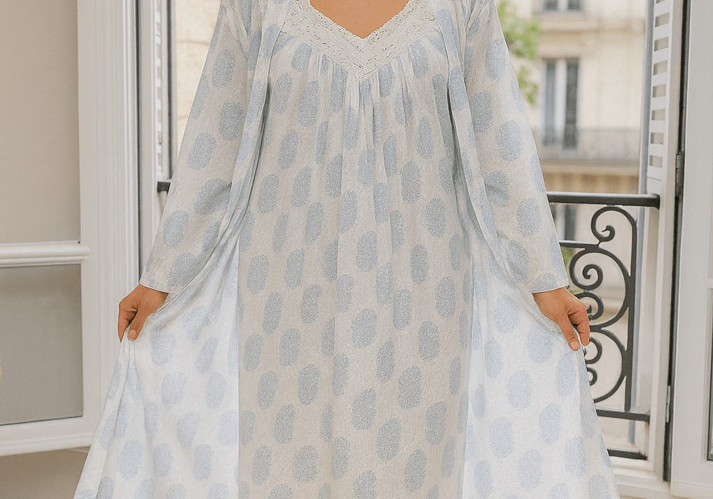 Woman wearing a white cotton robe with soft blue paisley print, standing in a sunlit room by a window.