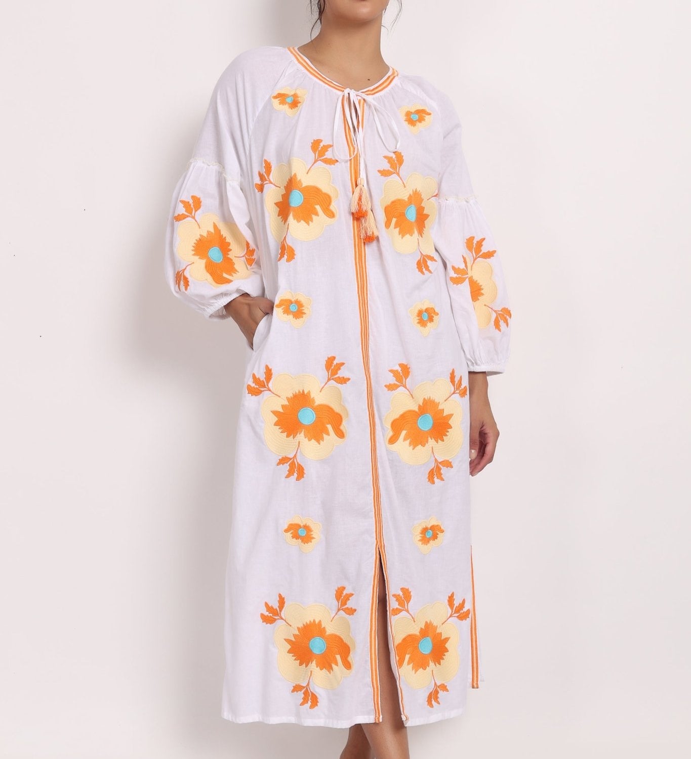 Model wearing white cotton sundress with beige floral embroidery, front view with tassel tie neckline.