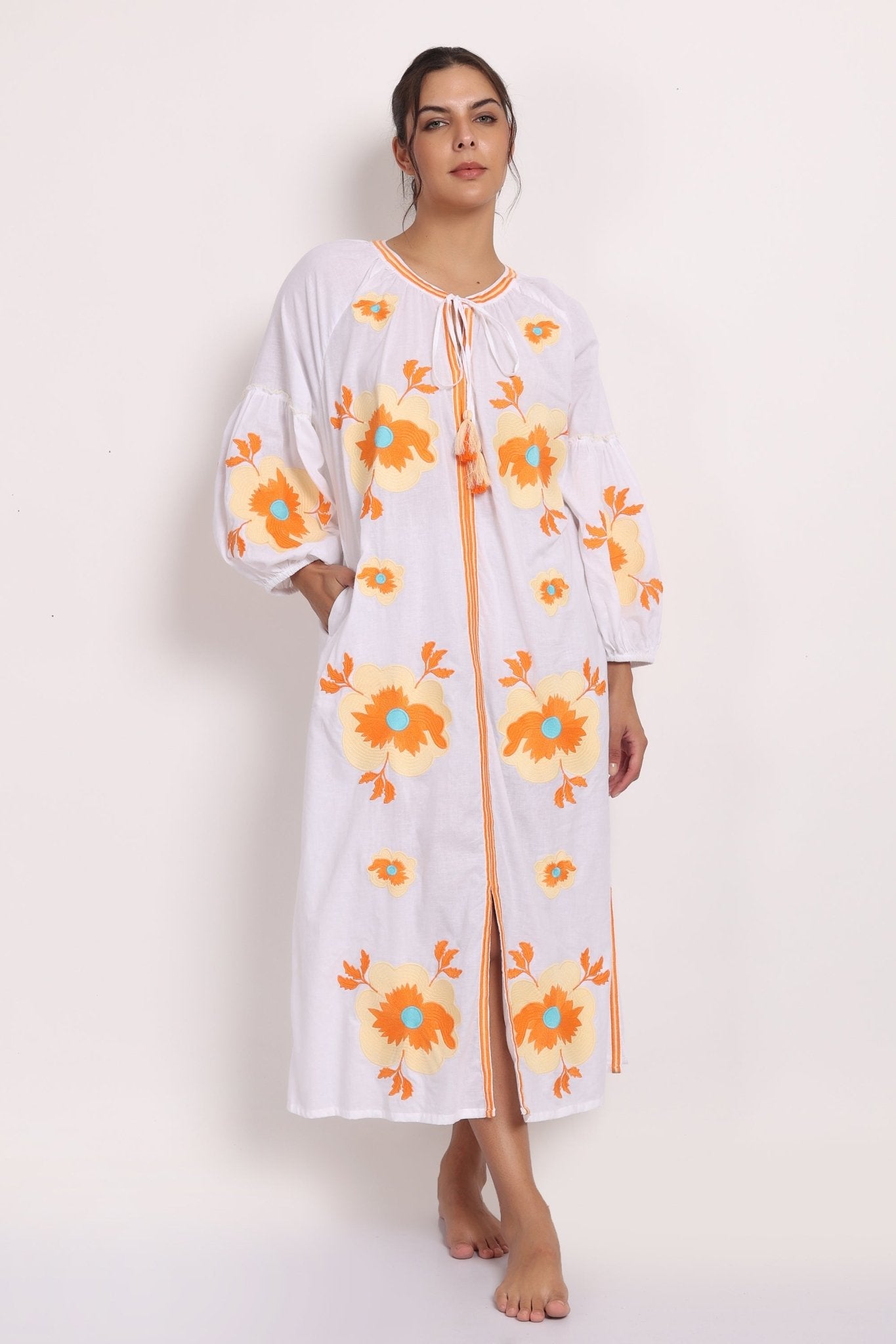 Model wearing white cotton sundress with beige floral embroidery, front view with tassel tie neckline.