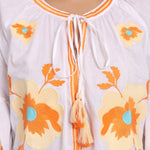 Close-up of beige floral embroidery and tassel neckline on white cotton dress.