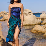 Woman in a blue and green dress standing on a rocky beach with clear blue water and sky.