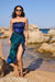 Woman in a blue and green dress standing on a rocky beach with clear blue water and sky.