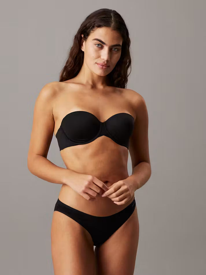 A woman modeling a black strapless bandeau bra and matching black underwear against a solid gray studio background.
