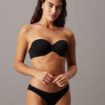 A woman modeling a black strapless bandeau bra and matching black underwear against a solid gray studio background.