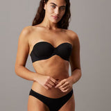 A woman modeling a black strapless bandeau bra and matching black underwear against a solid gray studio background.