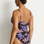 A woman modeling a one-piece swimsuit from the back, showing a black base with a vibrant pink, purple, and blue tropical floral pattern, set against a plain light gray studio background.