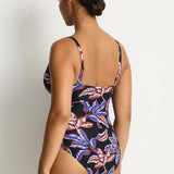 A woman modeling a one-piece swimsuit from the back, showing a black base with a vibrant pink, purple, and blue tropical floral pattern, set against a plain light gray studio background.