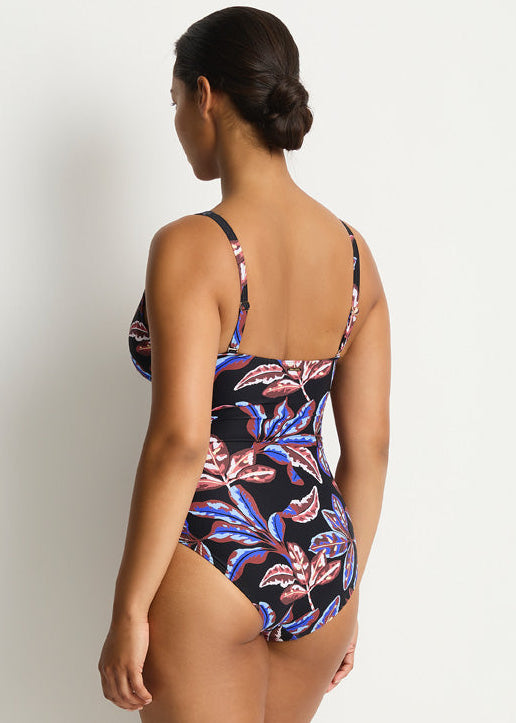 A woman modeling a one-piece swimsuit from the back, showing a black base with a vibrant pink, purple, and blue tropical floral pattern, set against a plain light gray studio background.