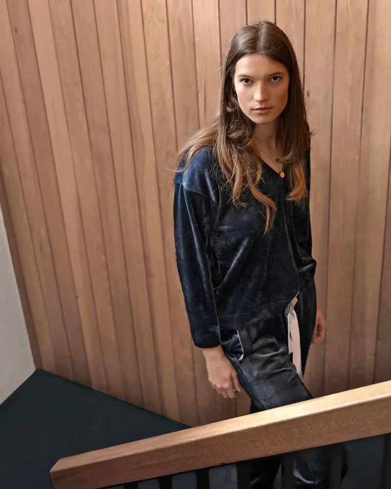 Woman in a dark blue velvet outfit standing against a wooden paneled wall.